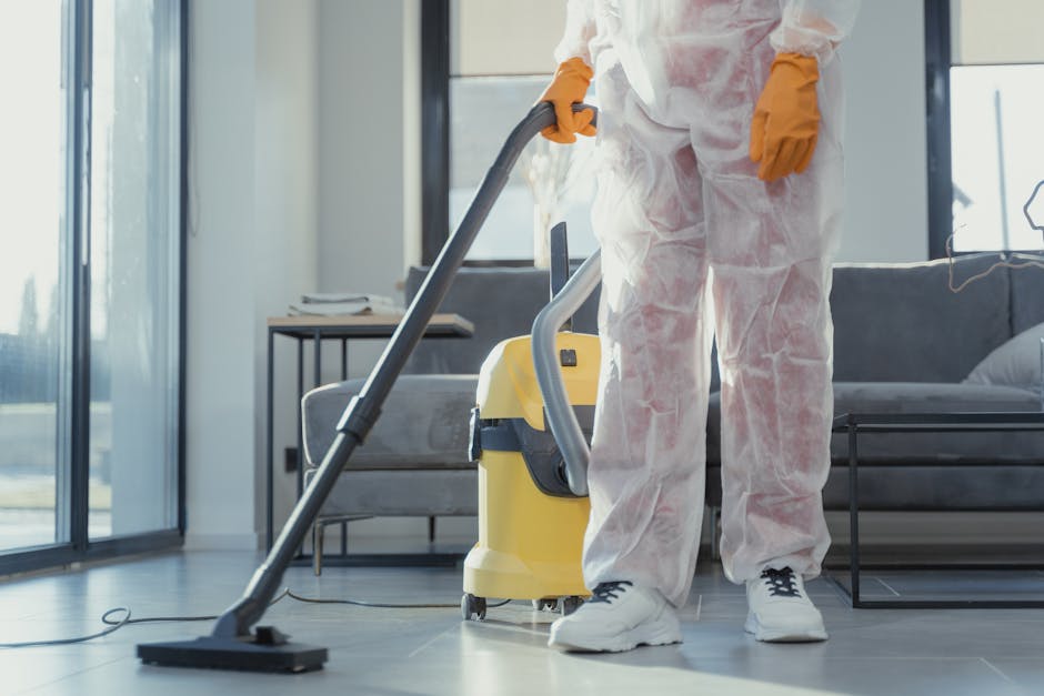 A professional cleaner wearing protective overalls and orange rubber gloves operates a yellow and gray vacuum cleaner on a tiled living room floor. The room features large glass sliding doors allowing