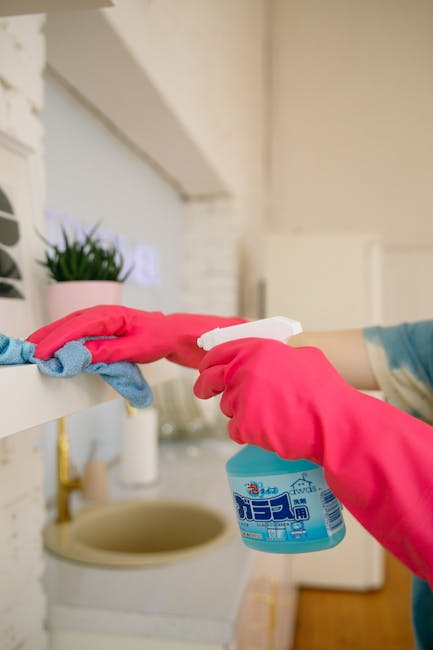 A professional cleaner wearing protective overalls and orange rubber gloves operates a yellow and gray vacuum cleaner on a tiled living room floor. The room features large glass sliding doors allowing